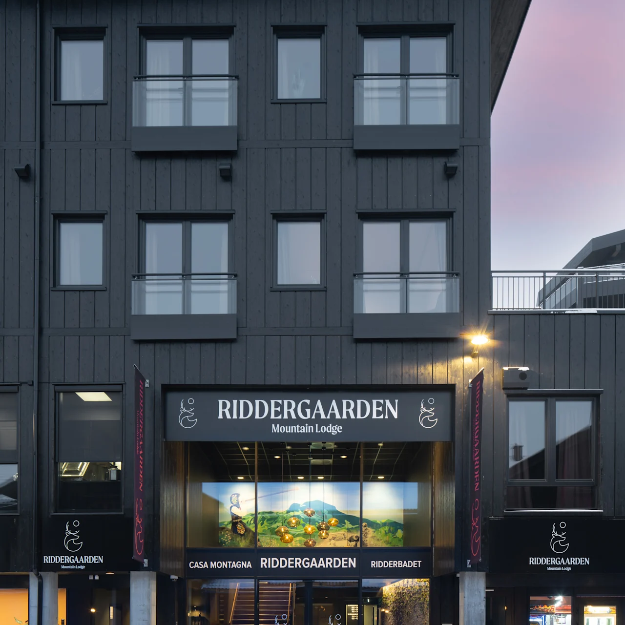 En flerfaglig mørk bygning med en belyst indgang læser "RIDDERGAARDEN Mountain Lodge." Snebeklædte omgivelser indikerer en vintersætning. Yderligere tekst: "CASA MONTAGNA," "RIDDERBADET."