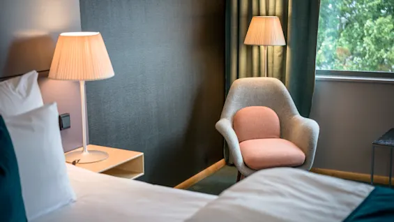 En hyggelig stol med lyserøde puder står ved en tændt lampe, ved siden af en seng med hvidt sengetøj og et natbord, i et blødt oplyst rum med grønne gardiner nær et vindue.