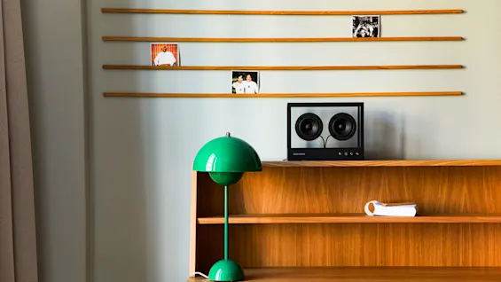 Et træbord holder en grøn lampe og en højttaler mærket "TRANSPARENT." Ovenfor er tre fotografier vist på vægmonterede træstave mod en lys væg.