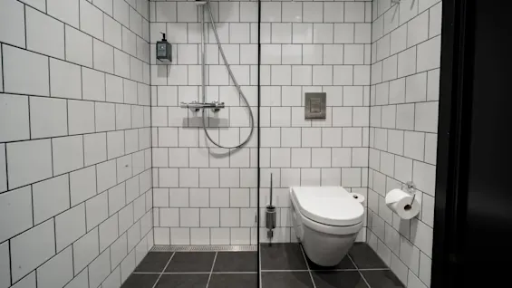 Nykyaikaisessa kylpyhuoneessa on seinään kiinnitetty wc-istuin ja suihku valkoisia kaakeleita vasten. Harmaat lattialaatat korostavat puhdasta, minimalistista tyyliä. WC-paperiteline on asennettu lähelle.