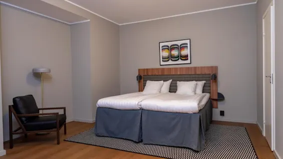 A bed with white sheets stands centered in a hotel room, accompanied by a black chair and a floor lamp. A colorful artwork decorates the wall above the bed.