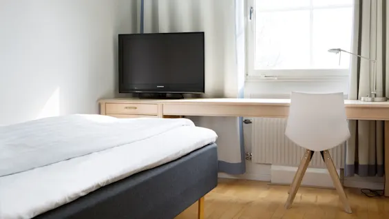 A television sits on a wooden desk in a bedroom; next to it, a white chair faces a window with drawn curtains. The bed is neatly made with white linens. The TV brand is "Thomson."