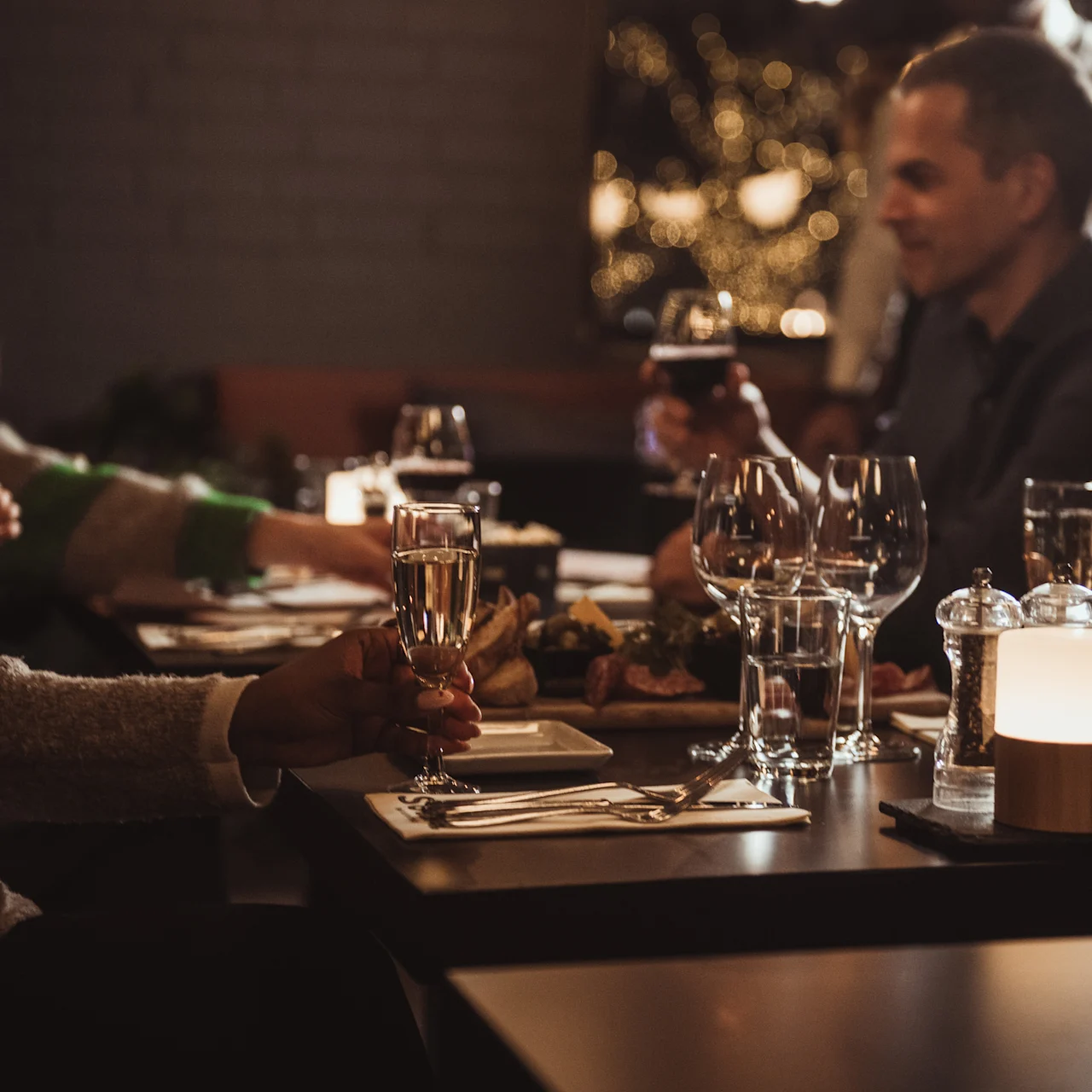People sit around a dimly lit restaurant table, holding glasses and engaging in conversation, with plates of food, utensils, and candles adding to the cozy atmosphere.