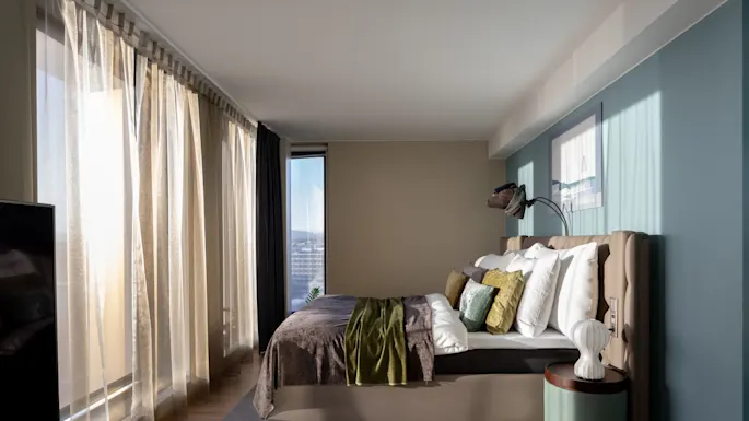 A neatly made bed with colorful pillows and a blanket sits beside a small lamp on a round table. Soft light filters through sheer curtains in a modern bedroom.