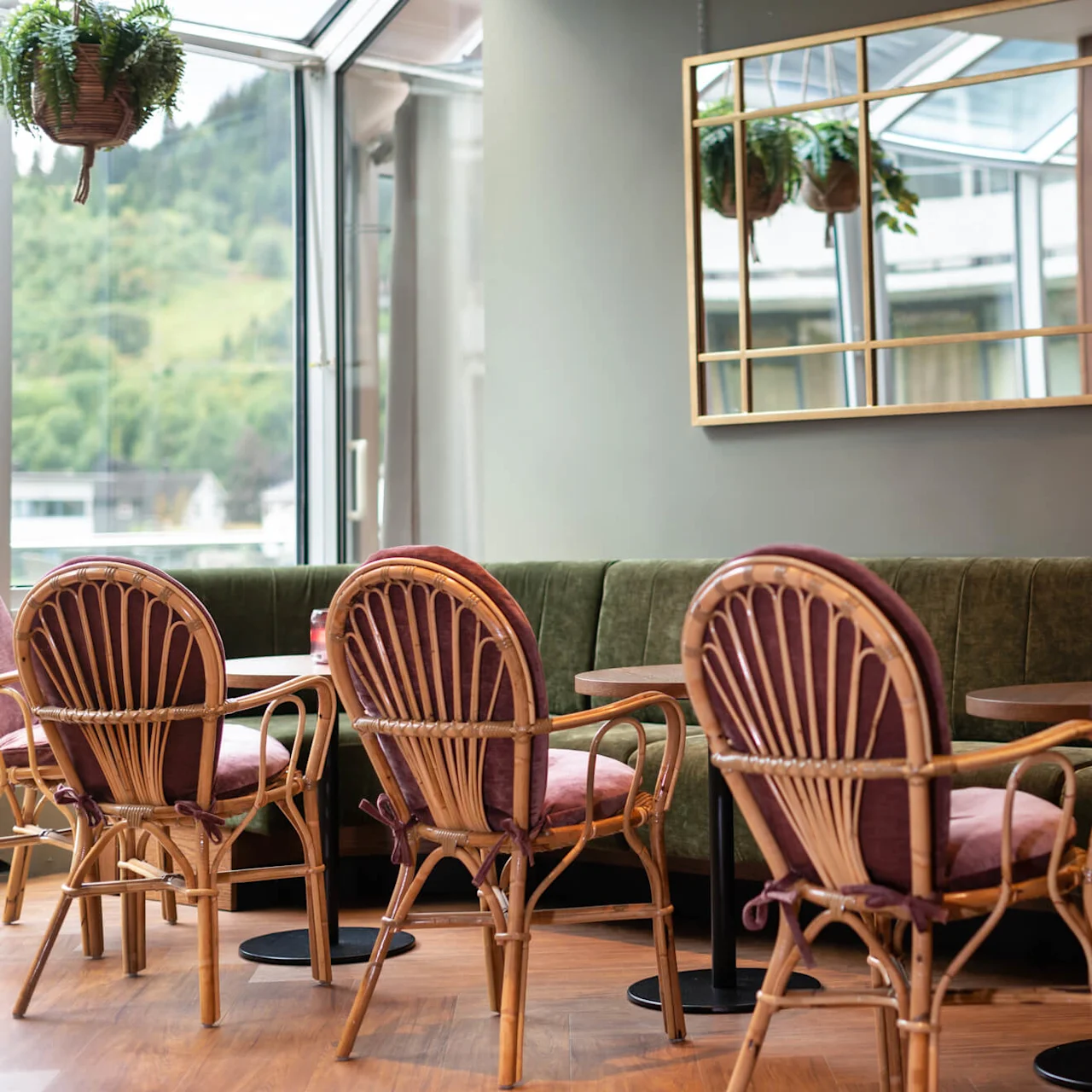 Trästolar med tygsitsar är placerade runt runda bord, varje med ett litet ljus, i ett mysigt café med gröna sammetssoffor och stora speglar som reflekterar hängande växter.