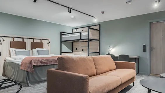 Et moderne hotellrom med en stor seng, køyesenger, en komfortabel sofa og en arbeidsplass.