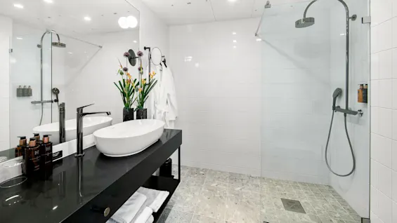 Modern bathroom with vessel sink on black countertop, glass shower, and white tiles. Robes hang on the wall, and flowers decorate beside toiletries. Towels are neatly stored below.