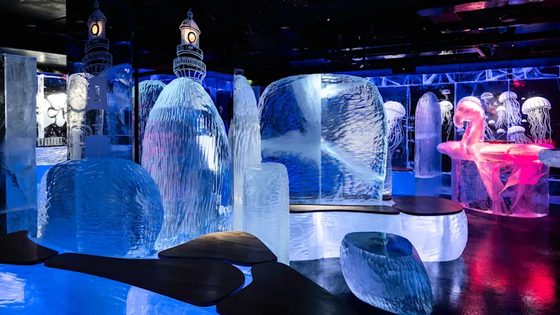 Ice sculptures are illuminated in blue and pink, featuring a flamingo and jellyfish designs. Wooden benches are placed amidst the icy structures in a dimly lit environment.