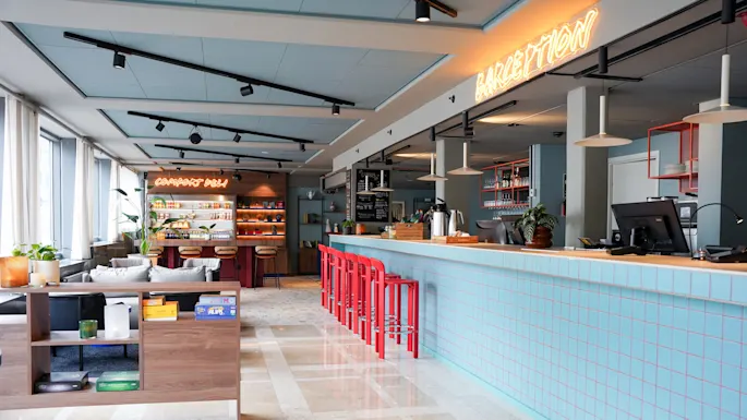 En lys café med en lang, blå flisebelagt disk med røde stole og neonlys, hvor der står "COMFORT DELI" og "BARCEPTION" i en hyggelig, moderne indretning.