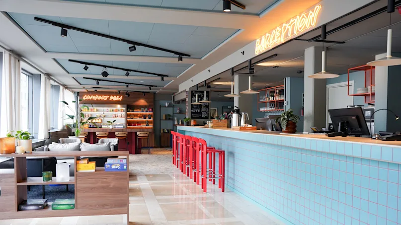 A brightly lit café features a long, blue-tiled counter with red stools and neon signs reading "COMFORT DELI" and "BARCEPTION" amidst a cozy, modern décor.