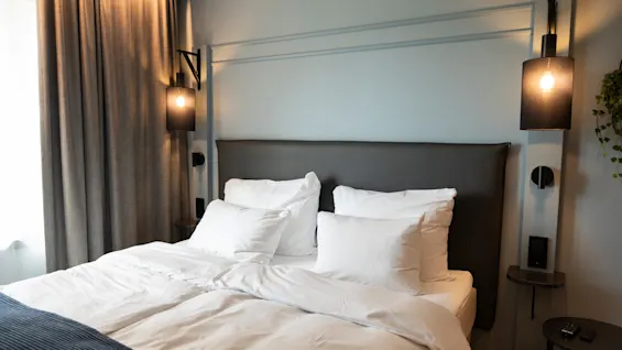 Et koselig hotellrom med en pent oppredd seng med hvitt sengetøy, en mørk sengegavl og stilige nattbordslamper.