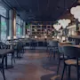 Dining tables and chairs sit orderly in a modern, stylish restaurant, illuminated by spherical pendant lights. Large windows provide natural lighting with a view of greenery outside. No text visible.