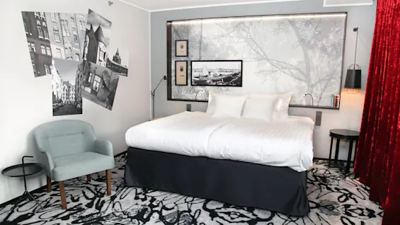 A neatly made bed with white linen in a bedroom with artistic monochrome wall decor, red curtains, and modern furniture. 