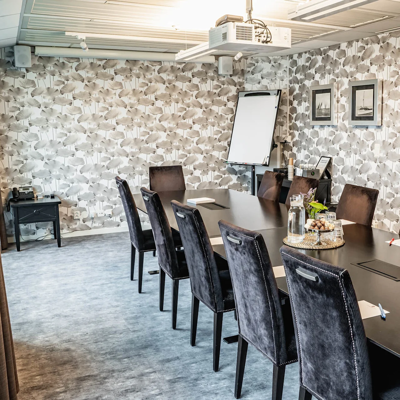 A long black conference table is surrounded by dark chairs, set with notepads, pens, and water bottles. The room features a patterned wallpaper and a flip chart.