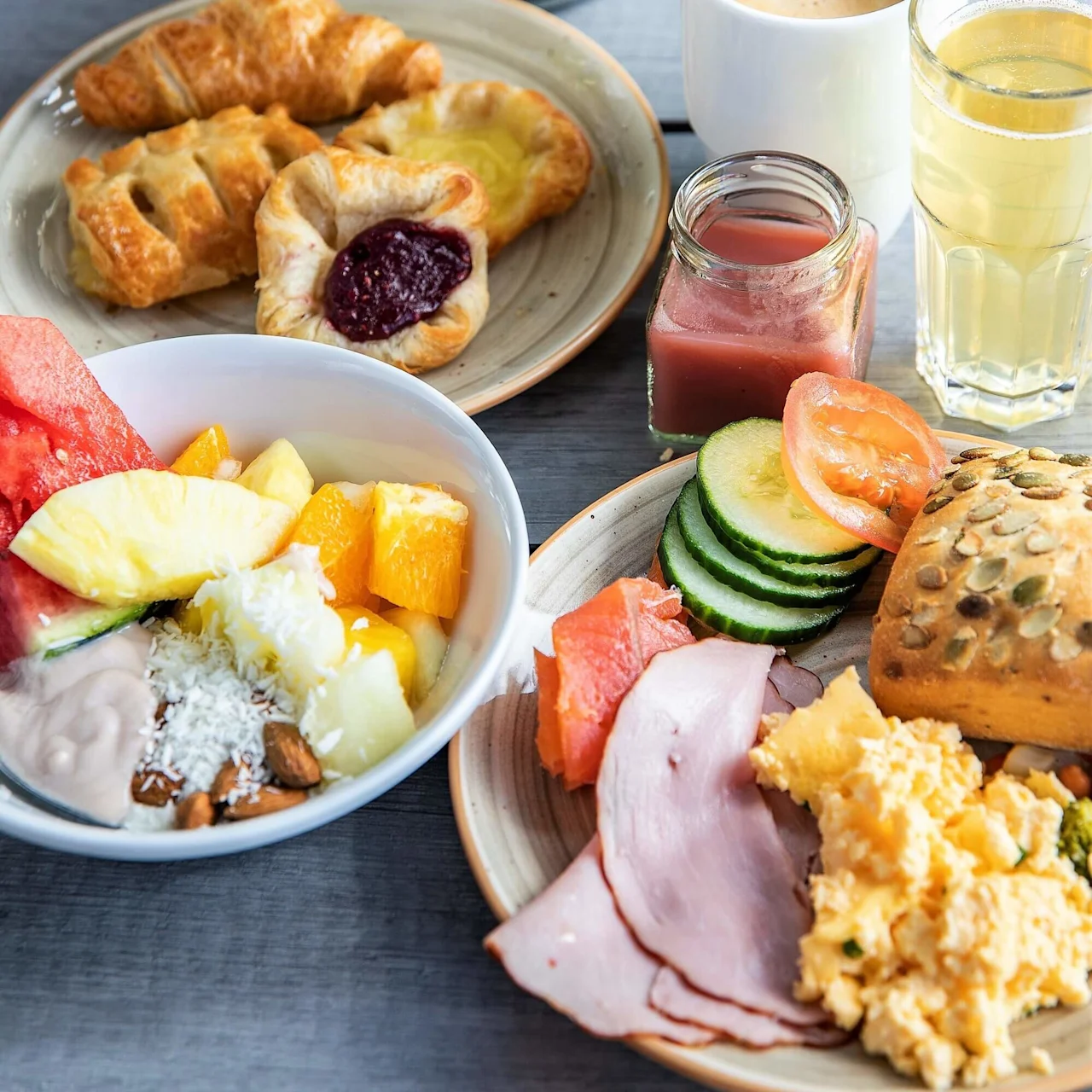Et variert frokostbord med bakverk, fruktsalat, kjøtt, egg og brød på tallerkener; ledsaget av kaffe, juice og en vase med hvite blomster på et trebord.