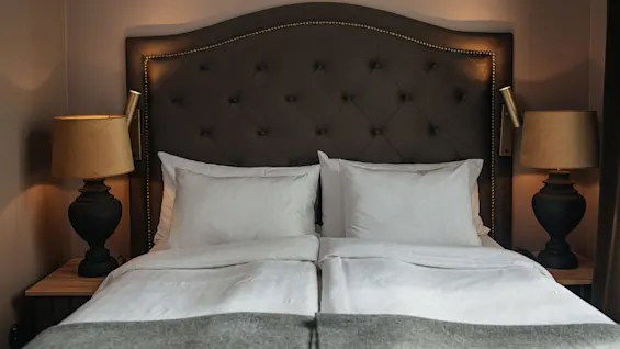 A neatly made double bed with white pillows and sheets stands between two dark bedside lamps on wooden tables, framed by a tufted, dark headboard and soft lighting. 