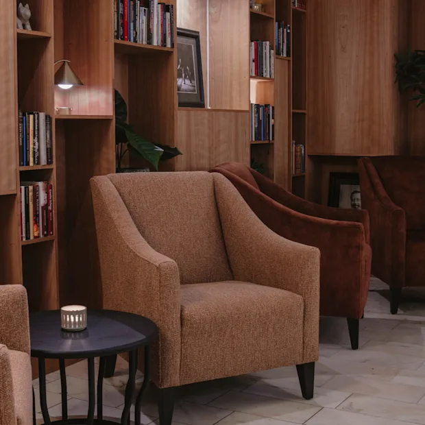 En varm och inbjudande hotellounge med bekväma sittplatser, bokhyllor och mjuk belysning. En bok med titeln Playing Cards är synlig.