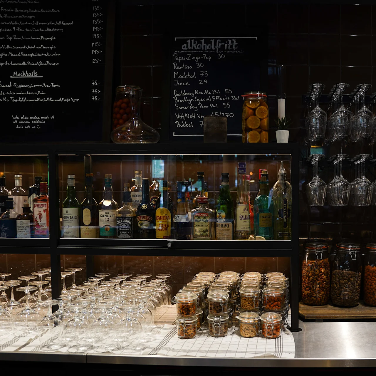 An array of alcohol bottles displayed behind a bar with stacked glasses and jars of snacks, below a blackboard menu.