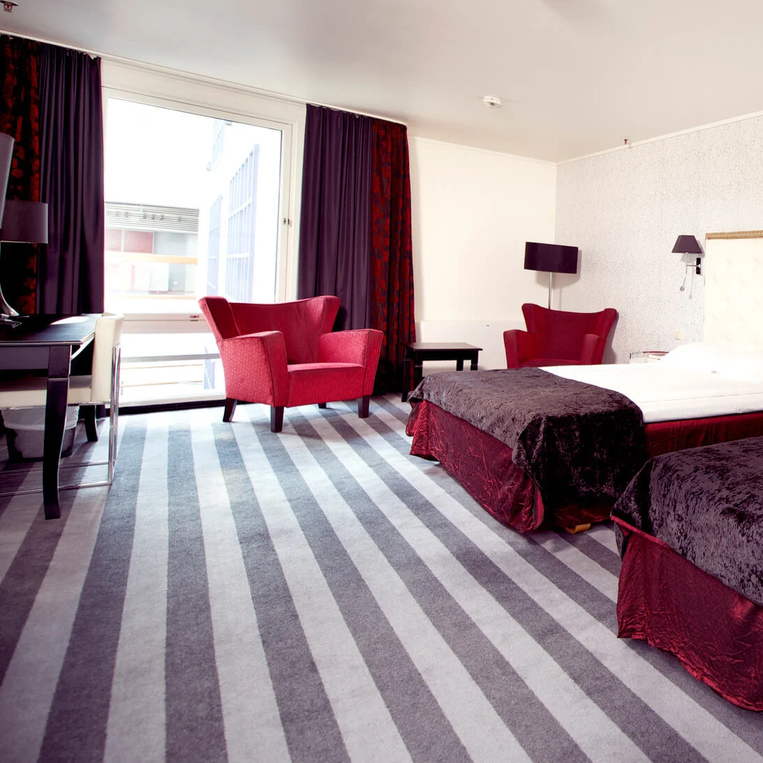 A hotel room with two beds, red chairs, and a balcony, illuminated by natural light.