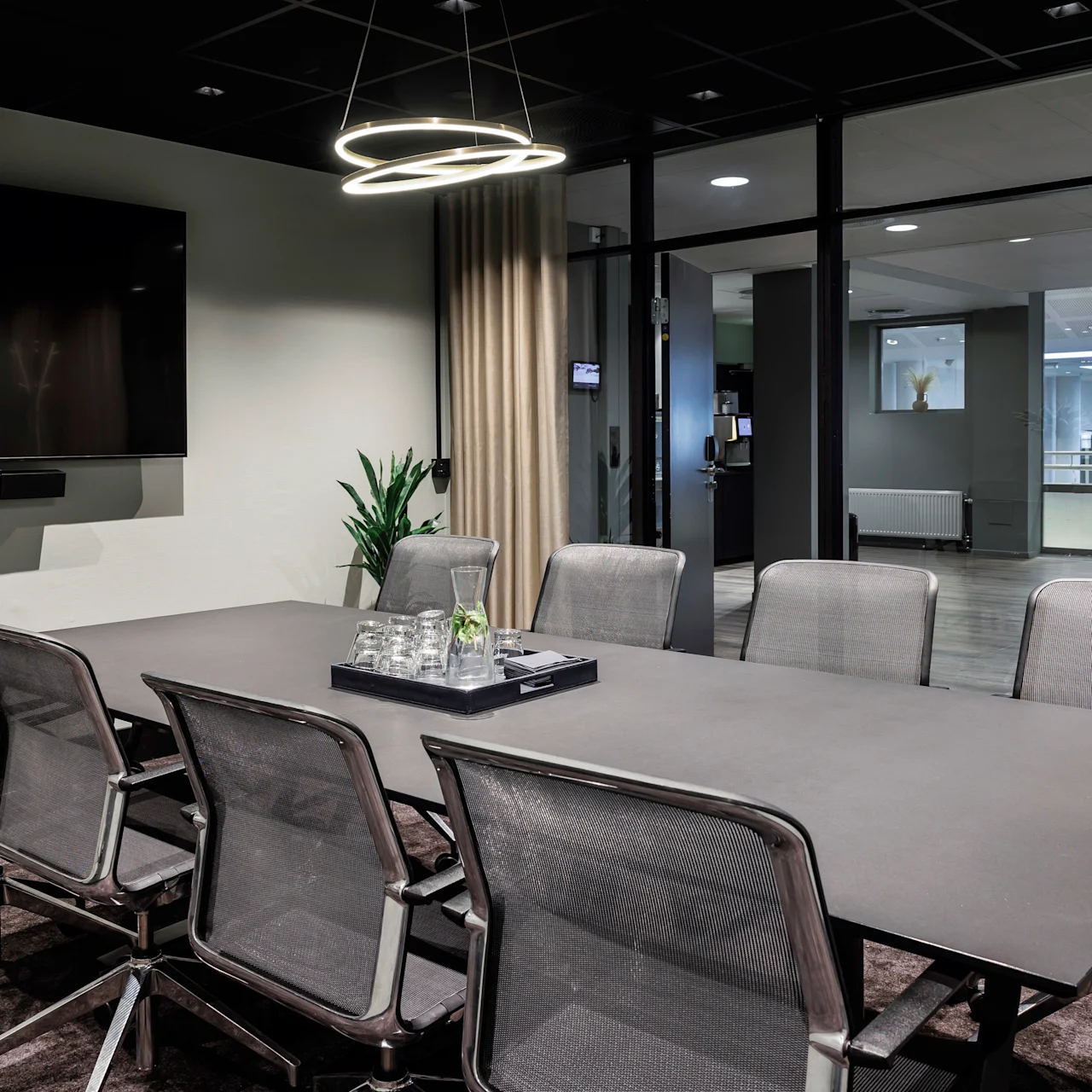 A conference table with eight chairs, under modern circular lights, sits in a meeting room with a TV on the wall and glass walls revealing an office environment outside.  