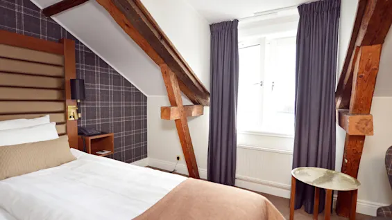 A cozy room with a neatly made bed, checked wallpaper, exposed wooden beams, a window, and minimalistic furnishings.