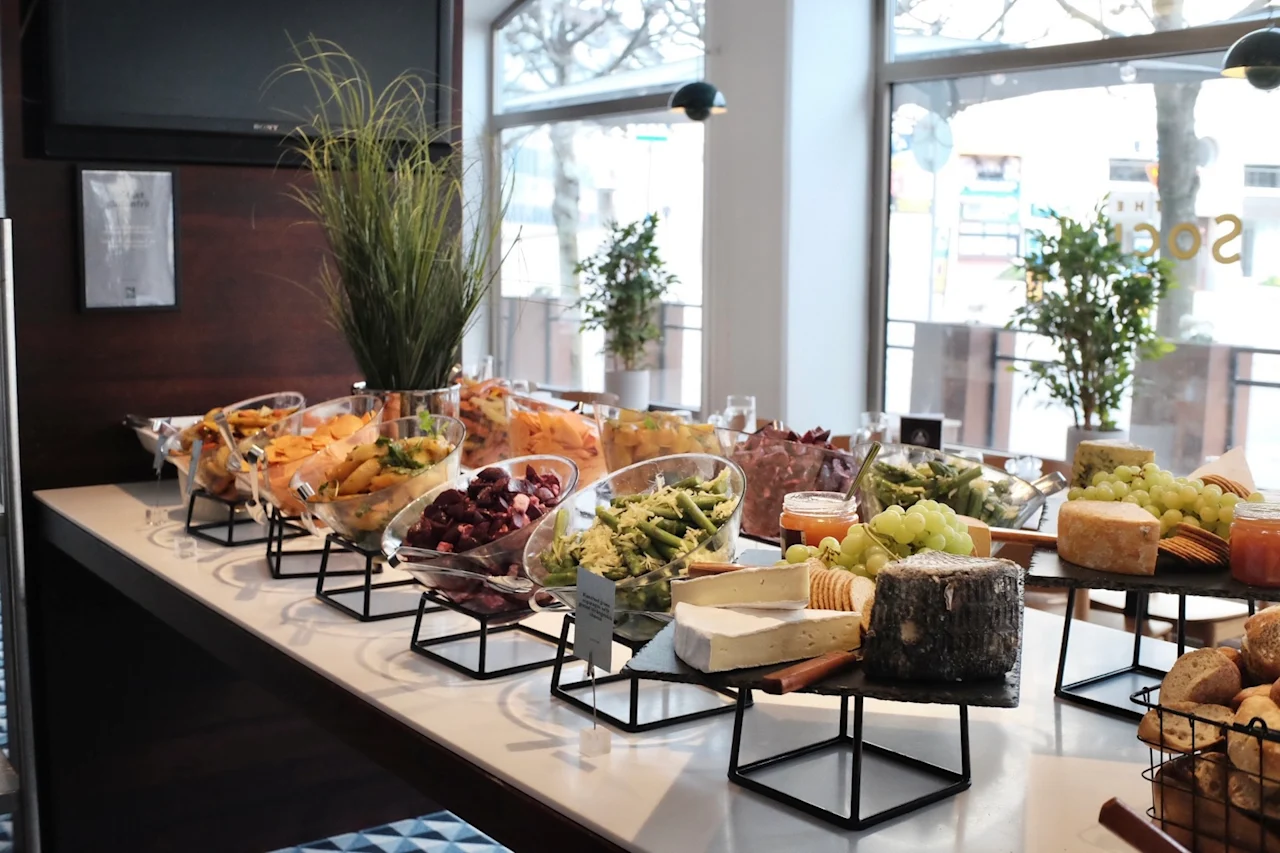 A buffet table presents a variety of colorful fruits and cheeses, neatly arranged in bowls and stands, set in a bright room with large windows and decorative plants. 