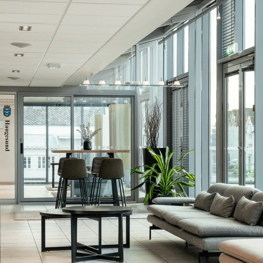 Ett modernt kontorsloungeområde har en grå soffa, ett runt soffbord och höga stolar nära en glasdörr med texten "Haugesund". Skyltar visar "Finnøy" och "Remesøy". Naturligt ljus strömmar genom stora fönster.