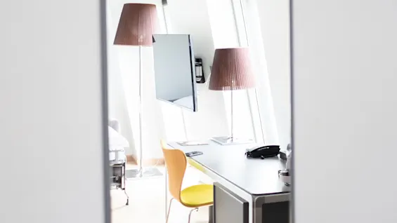 En väggspegel speglar ett modernt kontor med ett skrivbord och en gul stol, en monterad TV och golvlampor mot stora fönster i ett ljust rum.