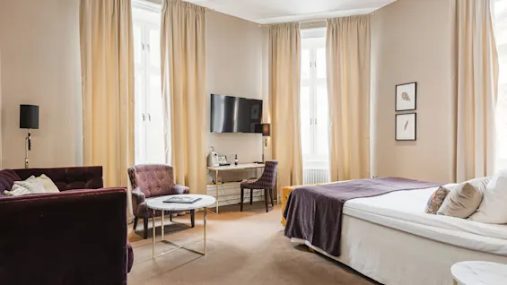 Elegant hotellrum med plyschsoffa, stol, skrivbord och säng i en neutral färgsättning.