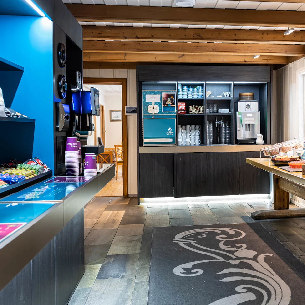 Et selvbetjent snackområde med kaffemaskin, snacks på en blå hylle og matbrett på et trebord. Godt opplyst rom med synlige trebjelker.