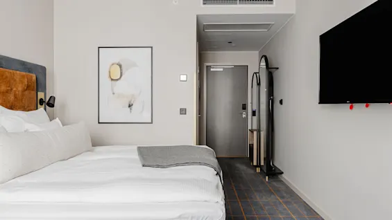En pænt redt seng med hvidt sengetøj og gråt tæppe, ved siden af en mørk TV-skærm, i et moderne, minimalistisk hotelværelse med abstrakt vægkunst og korridorudsigt.