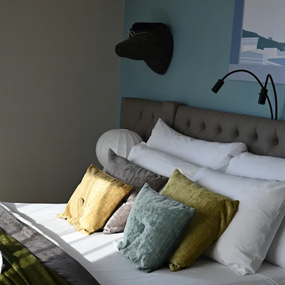Solljusbelyst hotellsäng med mjuka vita kuddar, färgglada prydnadskuddar, en stoppad grå sänggavel och en blå vägg.