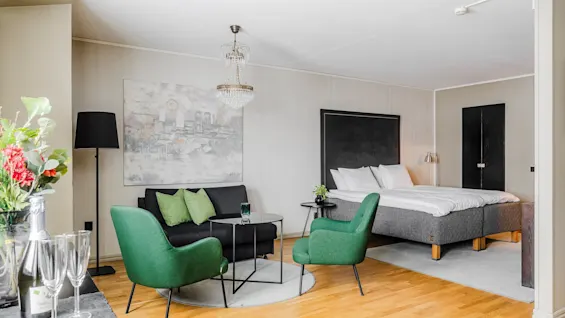 Ett mysigt hotellrum med en säng med vita sängkläder och svart huvudgavel, en sittgrupp med gröna stolar, ett litet bord och blommiga accenter under en ljuskrona.
