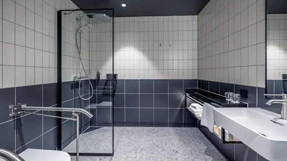 Bruseområde med glasindhegning, ved siden af en flisebelagt væg; bordplade med vask og håndklæder nedenunder, i et moderne badeværelsesmiljø.