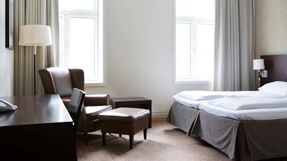 Hotellihuoneessa on suuri sänky valkoisilla vuodevaatteilla ruskean nojatuolin ja rahin vieressä. Pöytä ja tuoli täydentävät tilan, jota valaisee luonnonvalo kahdesta ikkunasta.