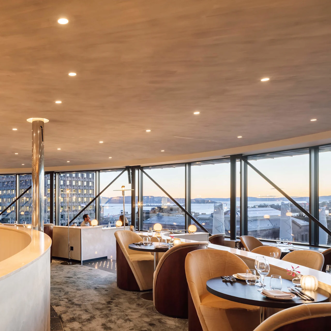 Moderne restaurantinteriør med panoramavinduer, der vender ud mod en havnefront i skumringen, med elegant møblering og blødt lys.