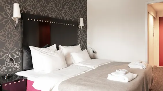 Viihtyisä hotellihuone, jossa on suuri sänky, valkoiset liinavaatteet ja tyylikäs kuvioitu tehosteseinä, kutsuen viihtyisään oleskeluun.