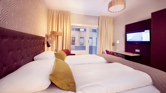 En pent oppredd seng med mykt hodegjerde i et moderne hotellrom, vinduer som viser en urban utsikt, et skrivebord og en flatskjerm-TV på veggen.