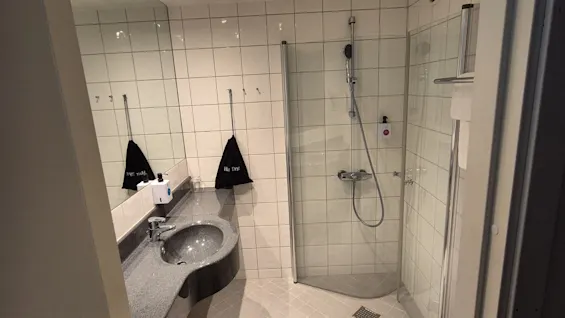 Bathroom with a gray countertop and sink next to a glass shower. White tiles cover walls and floor. Text on bags: "Rica Trysil." Ceiling lights illuminate the space.