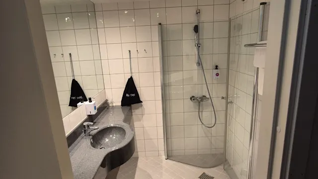 Kylpyhuone, jossa harmaa työtaso ja pesuallas, vieressä lasisuihku. Valkoiset laatat peittävät seinät ja lattian. Teksti pusseissa: "Rica Trysil." Kattovalot valaisevat tilaa.