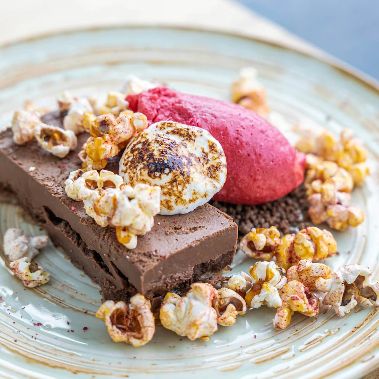 Chokladdessert garnerad med rostad marshmallow, bärsorbet och popcorn, arrangerad på en ljusblå tallrik på ett träbord.