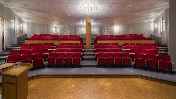 A wooden podium stands on a stage with a microphone, facing red auditorium seats in a bright lecture hall with a white ceiling.