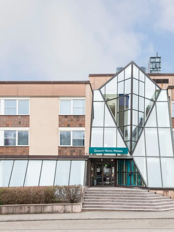 En modern hotellentré med glasfasad och geometrisk design, märkt "Quality Hotel Prisma," omgiven av buskar och ett stenbelagt område under en delvis molnig himmel.