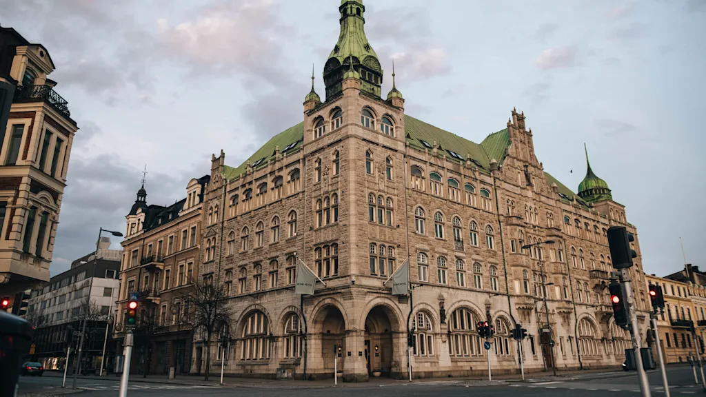 A large, ornate stone building with a green roof stands on a street corner. Surrounding it are other historic buildings and traffic lights. The sky is cloudy, casting a warm light.