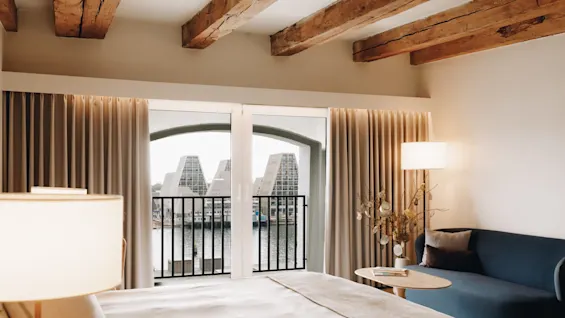 Mysigt hotellrum med takbjälkar i trä, en blå sofa och en balkong med utsikt över en kanal.