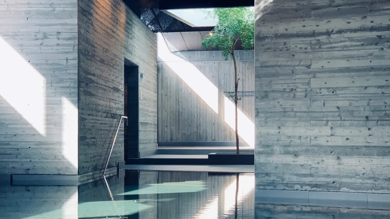 Indoor swimming pool reflecting sunlight, with shadows and a visible skylight, surrounded by concrete walls.