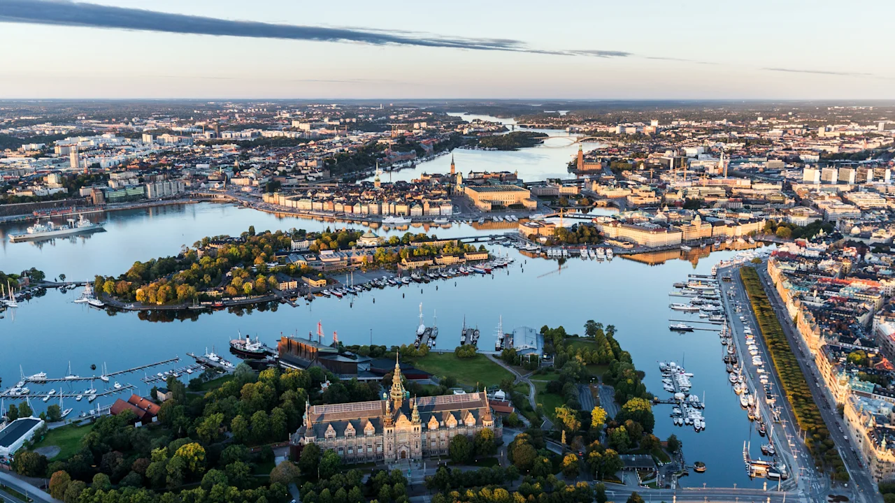 En historisk bygning er omgitt av frodige trær og vann, plassert i et urbant landskap. Mange båter ligger til kai langs vannkanten under en vidstrakt, klar himmel.