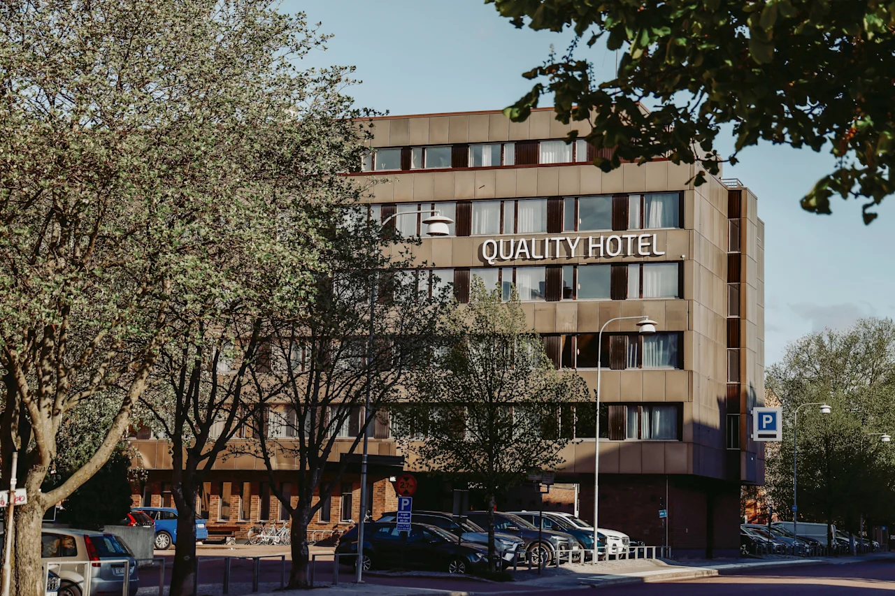 Quality Hotel sign on a modern, multi-story building surrounded by trees and parked cars. The trees are partly obscuring the view of the hotel, creating a natural urban setting.