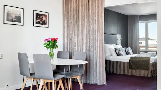 A dining area with a table set for four and a bedroom with an unmade bed, separated by a curtain, in a modern apartment.