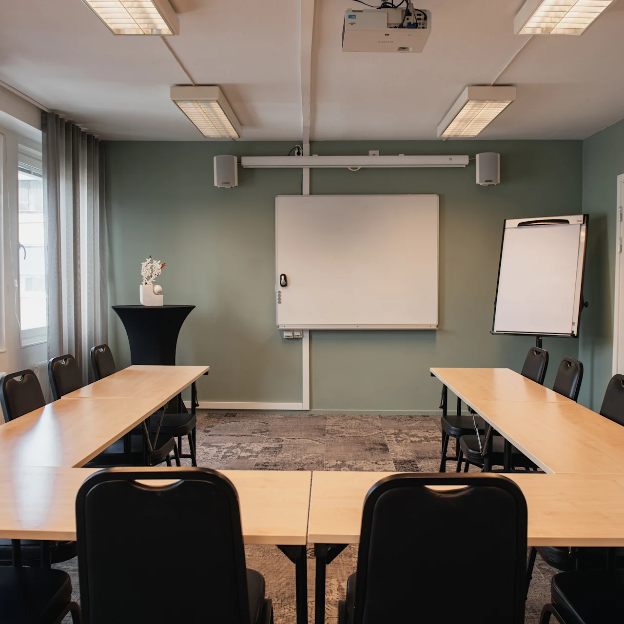 Conference room with a U-shaped arrangement of tables and chairs. A whiteboard and flip chart stand against a green wall. Overhead lights illuminate the space.